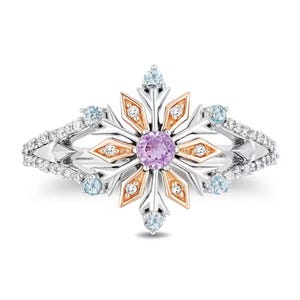 May include: A silver ring with a snowflake design. The center features a round purple gemstone, surrounded by gold and silver accents. Small blue gemstones and clear stones are set around the ring's band.
