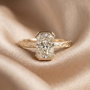 May include: A gold ring featuring a rectangular radiant-cut center stone set in a bezel with milgrain detailing. The band displays an engraved wheat-like pattern along the shank, providing a textured appearance to the metal surface.