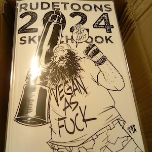 May include: A black and white sketchbook labelled "RUDETOONS 2024 SKETCHBOOK" with a cartoon illustration of a bearded person holding a large object. The person's shirt reads "VEGAN AS FUCK".