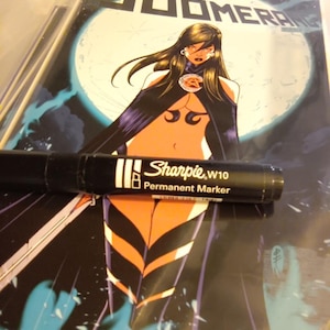 May include: A black Sharpie W10 permanent marker resting horizontally across a comic book titled Boomerang. The comic cover features a central illustration of a character with long dark hair, wearing a black cape and outfit against a large moon.