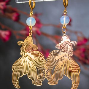 May include: Gold-colored fish earrings with intricate cut-out details. Each earring features a fish pendant suspended below a small, round, opalescent bead and a gold hoop. The earrings are displayed against a blurred background with pink and red accents.