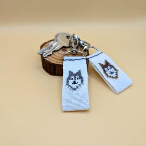 Könnte beinhalten: Zwei Schlüsselanhänger mit weißem Hintergrund und einem gestickten Husky-Hund-Design. Die Schlüsselanhänger sind an einem silbernen Schlüsselring befestigt. Die Schlüsselanhänger befinden sich neben einer Holzscheibe mit Schlüsseln.