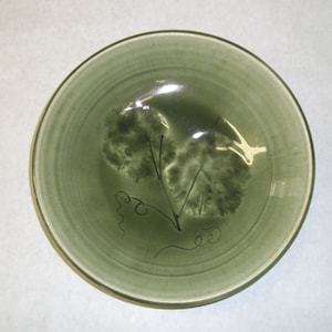 May include: A green ceramic bowl with a stylized design of leaves and vines. The bowl has a glossy finish and a slightly raised rim.