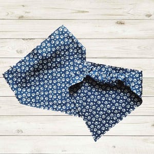 May include: Two blue pet bandanas with white paw prints on a light wood background. One bandana is rectangular, and the other is triangular. The bandanas are likely for dogs or cats.