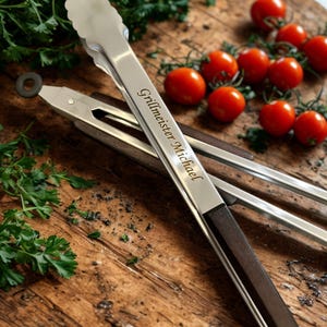 Könnte beinhalten: Grillzange aus Edelstahl mit dunklen Holzgriffen, auf einer Holzoberfläche mit Kirschtomaten und Petersilie. Auf der Zange steht der Schriftzug "Grillmeister Michael".
