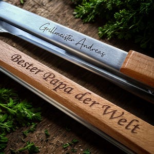 Könnte beinhalten: Nahaufnahme von zwei Grillzangen. Eine ist aus Edelstahl mit der Aufschrift "Grillmeister Andreas". Die andere ist aus Holz mit der Aufschrift "Bester Papa der Welt". Frische Kräuter sind verstreut.