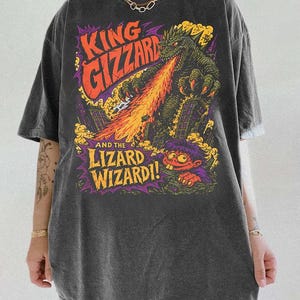 May include: A charcoal gray short-sleeve t-shirt featuring a graphic print of a green monster breathing fire over a city, with the text "KING GIZZARD AND THE LIZARD WIZARD!" in orange and yellow, and a small cartoon character at the bottom.