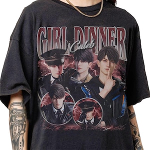 Può includere: Una t-shirt grigio antracite a maniche corte con una stampa grafica che riporta il testo "GIRL DINNER" e "Caleb" sopra quattro illustrazioni di un giovane uomo in vari abiti, su uno sfondo di fulmini rossi e nuvole scure.