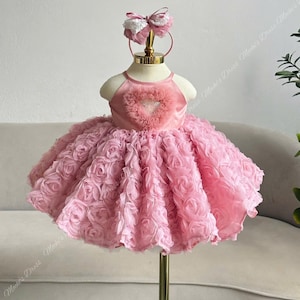 May include: A pink baby dress with a halter neckline and a heart-shaped embellishment on the bodice. The skirt is made of layered rosettes. A matching pink headband with a bow completes the outfit. The dress is displayed on a mannequin.