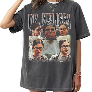May include: A charcoal grey short-sleeve t-shirt featuring a graphic print with the text DR. MELISSA KING. The design includes five repeated portraits of a person wearing glasses, arranged in a collage style with a lightning bolt background.