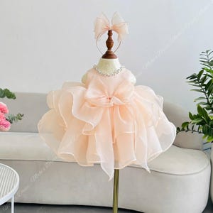 May include: A peach-coloured sleeveless dress featuring a voluminous, multi-layered organza skirt with ruffled edges. The bodice is adorned with a crystal-embellished neckline and a large central bow. Includes a matching peach organza hair bow.