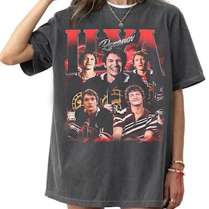 May include: A charcoal grey short-sleeve t-shirt featuring a large graphic print of a hockey player in various poses. The text "ILYA" is printed in large red letters at the top, with the name "Rozanov" in white script lettering centred above the images.
