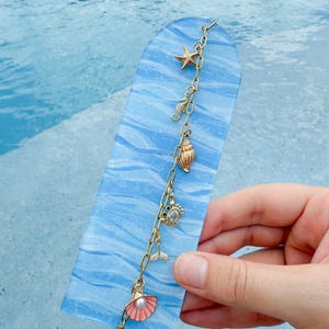 May include: A blue, arched bookmark with a gold chain featuring ocean-themed charms. Charms include a starfish, seashell, seahorse, and a mermaid tail. A pink shell charm with a pearl hangs at the bottom.