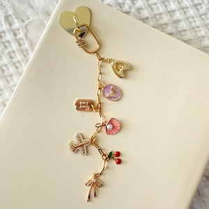 May include: A gold-colored charm bracelet with various charms, including a heart, a key, a shell, a plane, cherries, and a bow. The charms are attached to a chain and are resting on a cream-colored surface.
