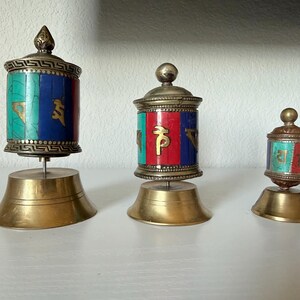 May include: Three decorative prayer wheels with brass bases. The wheels are cylindrical, with colorful inlays of turquoise, red, and blue. Each wheel has gold accents and a decorative top. The wheels are of varying sizes, with the largest approximately 5 inches tall.
