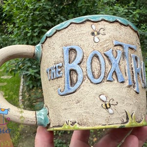 May include: A ceramic mug with the text "THE BOXTR" in blue lettering. The mug has a beige background with bee and grass details. The handle is decorated with a blue horse and floral design.