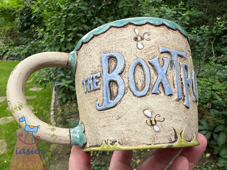 May include: A ceramic mug with the text "THE BOXTR" in blue lettering. The mug has a beige background with bee and grass details. The handle is decorated with a blue horse and floral design.