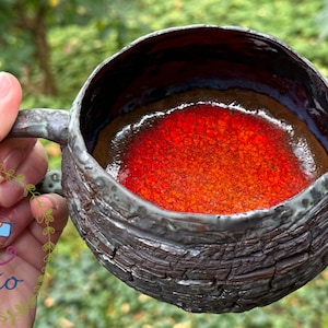 May include: A ceramic mug with a dark, textured, bark-like exterior finish and a smooth, vibrant red interior glaze. The mug features a rounded shape, a sturdy handle, and a distinct contrast between the rough outer surface and the glossy red centre.