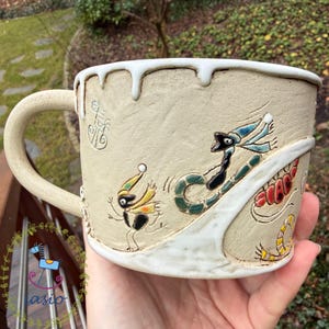 May include: A beige ceramic mug with a white drip glaze along the rim. The exterior features embossed, hand-painted illustrations of whimsical creatures in yellow, black, blue, and red, along with a raised white diagonal band across the surface.