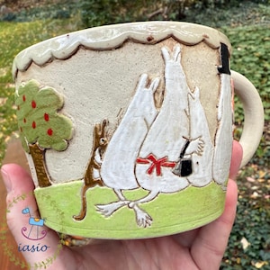 Könnte beinhalten: Cremefarbene Keramiktasse mit einem skurrilen Design. Die Tasse zeigt ein Relief von Cartoonfiguren, einen Baum mit roten Äpfeln und eine grüne, grasbewachsene Basis. Der Griff hat eine passende cremefarbene Farbe.