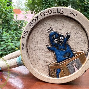 May include: A ceramic mug featuring a blue cartoon character emerging from a box. The mug has a beige background with the words "THE BOXTROLLS" around the rim. The handle is off-white with blue accents. The mug is on a wooden surface.