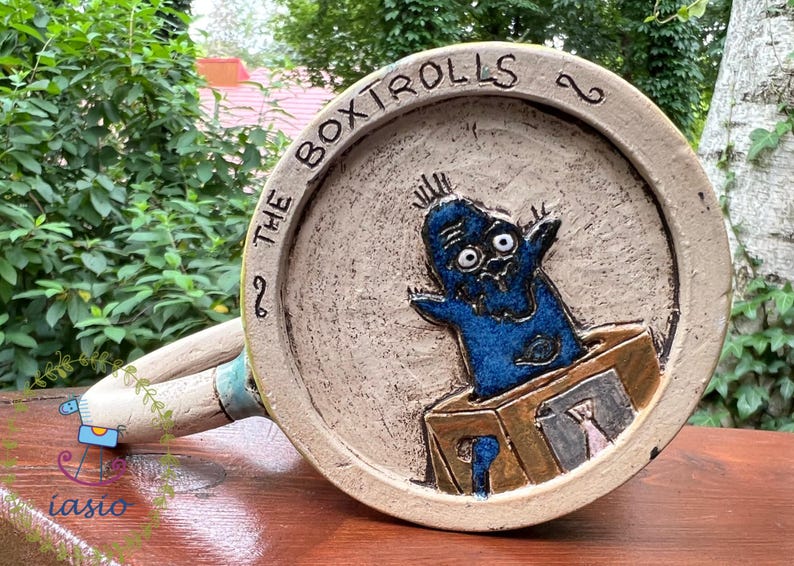May include: A ceramic mug featuring a blue cartoon character emerging from a box. The mug has a beige background with the words "THE BOXTROLLS" around the rim. The handle is off-white with blue accents. The mug is on a wooden surface.