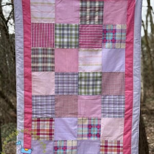 Puede incluir: Una colcha de retazos rectangular con una cuadrícula de cuadrados en varios tonos de rosa, lavanda y blanco. Los patrones de tela incluyen cuadros vichy, tartán y colores sólidos. La colcha está enmarcada por un borde rosa y un borde exterior lavanda.