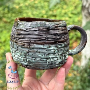 Może przedstawiać: Ceramiczny kubek o teksturowanej powierzchni przypominającej korę, z ciemnobrązową górną częścią oraz nakrapianą, turkusowo-czarną dolną częścią. Kubek ma zaokrąglony korpus i zakrzywione ucho, prezentując warstwowe, organiczne wykończenie.
