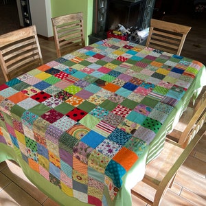 May include: A patchwork tablecloth with a green border, featuring a variety of colorful squares and patterns. The tablecloth is laid out on a wooden table with four chairs.