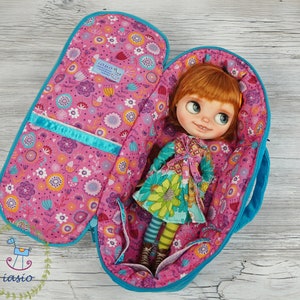 以下が含まれることがあります： ジッパー付きの青とピンクの花柄のトラベルケース。ケースが開いており、長い赤い髪と花柄のドレスを着た人形が入っています。