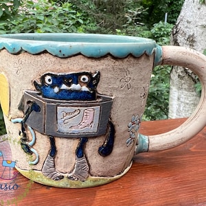May include: A handmade ceramic mug with a whimsical monster design. The mug features a blue monster figure with a box-shaped body and the word "Dames" with a boot illustration. The mug has a light blue rim and handle, with floral and star details.