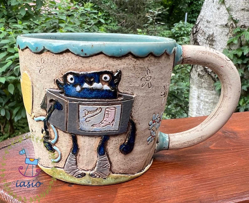 May include: A handmade ceramic mug with a whimsical monster design. The mug features a blue monster figure with a box-shaped body and the word "Dames" with a boot illustration. The mug has a light blue rim and handle, with floral and star details.