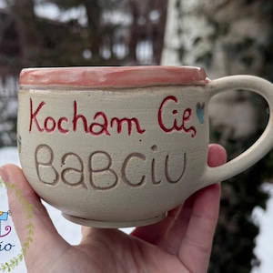 Può includere: Tazza in ceramica color crema con bordo rosa e manico grande. La tazza presenta scritte rosse che dicono "Kocham Cię, Babciu". Un piccolo logo con un cavallo a dondolo è visibile in basso a sinistra.