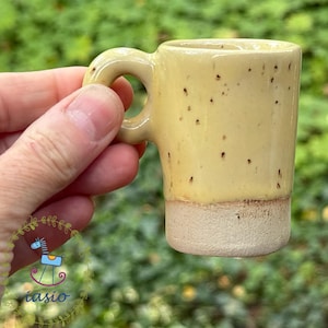 Könnte beinhalten: Ein kleiner Keramikbecher mit einem glänzenden, hellgelben oberen Teil mit dunkelbraunen Sprenkeln und einer matten, strukturierten beigen Basis. Der Becher hat einen abgerundeten Henkel an der Seite und einen zylindrischen Körper mit zweifarbigem Design.