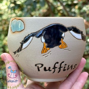 Può includere: Una ciotola in ceramica beige con un'illustrazione dipinta a mano di una pulcinella di mare bianca e nera in volo con zampe arancioni brillanti. Una piccola nuvola azzurra è dipinta sopra l'uccello e la parola Puffins è scritta in nero.