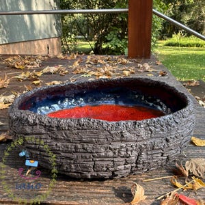 May include: A low, wide ceramic bowl with a dark, rough, bark-like exterior texture. The interior features a smooth, glossy finish with a deep blue rim that transitions into a vibrant, textured red centre, creating a striking contrast.