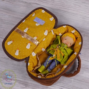 May include: A brown fabric travel case with a zipper closure and a yellow fabric lining with a snowman pattern. The case is open and a doll with blonde hair is inside wearing a green jacket and blue jeans.