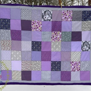 May include: A handmade patchwork quilt in shades of purple, lavender, and gray. The quilt features a variety of patterned squares, including polka dots, floral prints, and animal motifs. It is bordered with a dark purple fabric and hung outdoors.