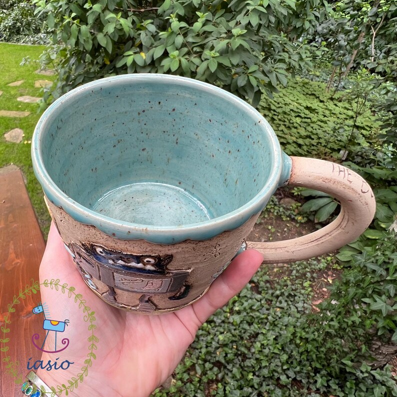 May include: Handmade ceramic mug with a light blue interior and a textured, tan exterior. The mug features a sculpted design and a large, rounded handle. The rim is a contrasting light blue. The handle has the word "THE" on it.