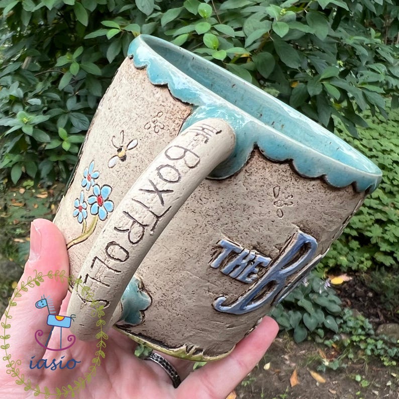 May include: A handmade ceramic mug with a light brown exterior and a turquoise rim and handle. The mug features the text "THE BOXTROLL" in a whimsical font, along with floral and insect designs. The mug is held in a person's hand.