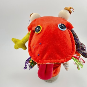 Op de afbeelding: Een rode pluche pop met een lange tong die eruit steekt. De pop heeft grote zwarte ogen met gele en blauwe knopen. De pop heeft een groen, geel en paars stoffen lichaam met een geruite stoffen sjaal.