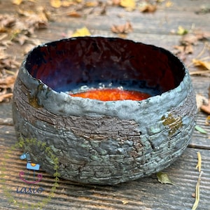 May include: A round ceramic bowl with a dark, textured, bark-like exterior in shades of gray and brown. The interior features a smooth, glossy finish with a deep red and orange center, contrasting with the dark, uneven, and rugged rim.