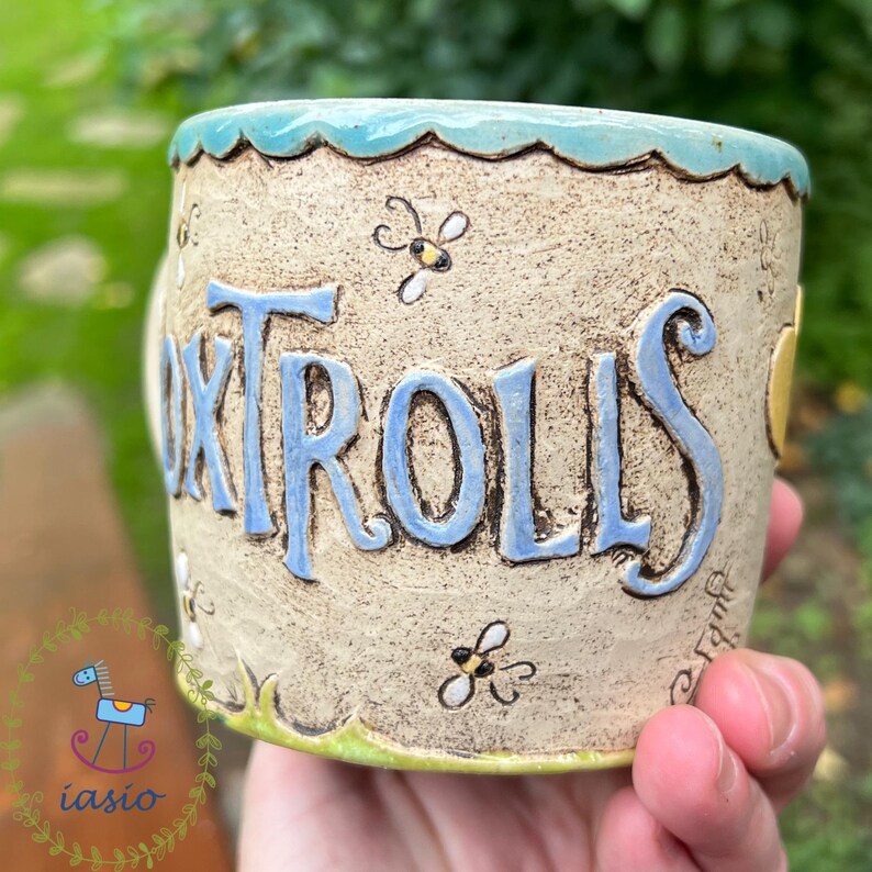 May include: A handmade ceramic mug with the word "ONTROLLS" in blue, raised letters. The mug has a light beige background with bee and floral designs. The rim is light blue, and the base has a green trim. The mug is held in a person's hand.