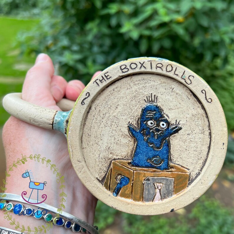 May include: A handmade ceramic mug featuring a blue cartoon character from "The Boxtrolls" movie. The mug has a beige background with the words "The Boxtrolls" around the rim. The handle is a light beige color.