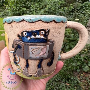 May include: Handmade ceramic mug with a whimsical monster design. The mug features a blue monster with large eyes and a box-like body. The mug's rim is scalloped and light blue. The handle is a light beige color. The mug has the word "Dame" on the front.