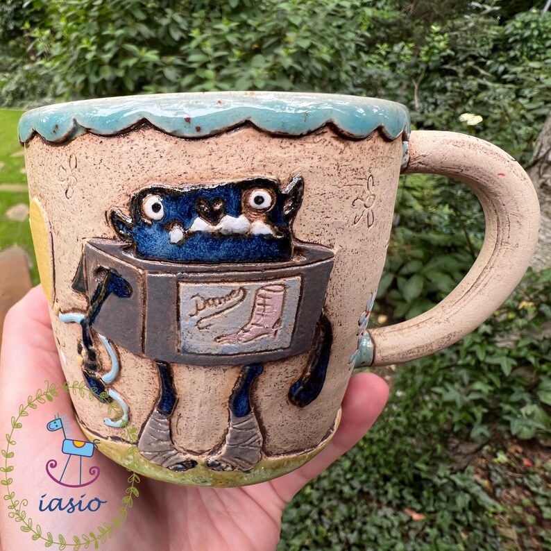 May include: Handmade ceramic mug with a whimsical monster design. The mug features a blue monster with large eyes and a box-like body. The mug's rim is scalloped and light blue. The handle is a light beige color. The mug has the word "Dame" on the front.