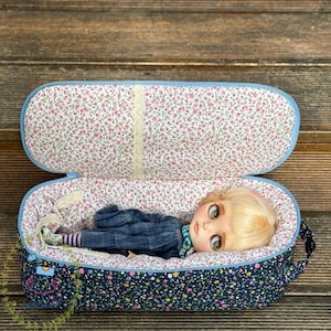 May include: An oval-shaped fabric carrying case with a blue zipper and a dark blue exterior featuring a colorful floral pattern. The interior is lined with a light-colored fabric with a small pink floral print, containing a doll with blonde hair.