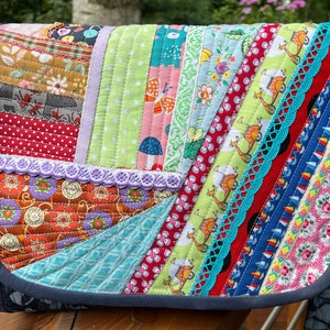 May include: A colorful patchwork messenger bag with a flap closure. The bag features a variety of patterned fabrics in red, green, blue, and orange, with decorative lace trim. The bag has a dark blue strap and trim.