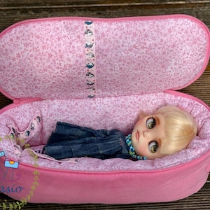 May include: A pink fabric carrying case with a floral interior lining and a central ribbon featuring small illustrations. Inside, a doll with blonde hair and large eyes wears a dark blue dress and a blue and green patterned neck accessory.