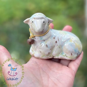 Puede incluir: Una pequeña figura de oveja de cerámica con un esmalte brillante multicolor en tonos azul pálido, amarillo y blanco, con motas marrones. La oveja lleva un cordón amarillo fino alrededor del cuello con una pequeña campana de metal marrón oscuro.